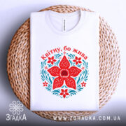 Футболка квітну бо жива, біла, складена на плетеній кошику, принт червоного квітки та зелених листків, фон білий, текст зверху червоним шрифтом.