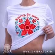 Футболка квітну бо жива, біла, лежить на поверхні, принт червоного квітки та зелених листків, фон світло-сірий, етикетка з зеленим шнурком.