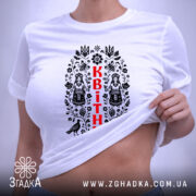 Футболка квітни білого кольору, складена на поверхні, фон світло-сірий, принт з червоним текстом 'КВІТНІ' та чорними малюнками, включаючи українські символи, з етикеткою на шнурку.
