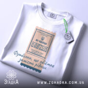 Футболка квиток, біла, на плоскій поверхні, фон світлий, принт у вигляді квитка з текстом, прикріплена етикетка з зеленим шнурком.