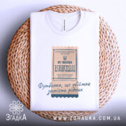 Футболка квиток, біла, на плоскій поверхні, фон світлий, принт у вигляді квитка з текстом, розташована на плетеній кошику.