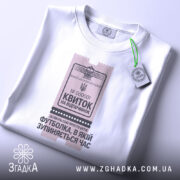 Футболка квиток на відпочинок, біла, знята зверху, фон світло-фіолетовий, принт у вигляді квитка з текстом 'Квиток на відпочинок', на комірці є етикетка.