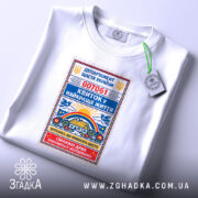 Футболка квиток у найкраще життя. Біла футболка, розкладена на поверхні, з принтом у вигляді квитка, що містить кольорові елементи. На футболці видно етикетку з логотипом. Фон світлий, з м'яким освітленням.