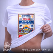 Футболка квиток у найкраще життя. Біла футболка, розкладена на поверхні, з принтом у вигляді квитка, що містить кольорові елементи. На футболці видно етикетку з логотипом. Фон світлий, з м'яким освітленням.