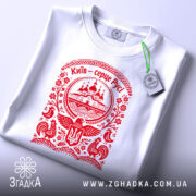 Футболка Київ серце Русі, біла, на плоскій поверхні, з червоним принтом, фон світло-сірий, принт з зображенням церкви та елементами українського орнаменту, з етикеткою.
