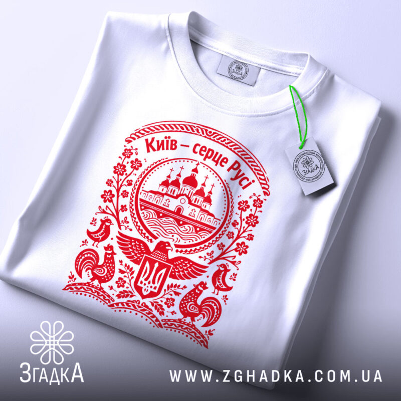 Футболка Київ серце Русі, біла, на плоскій поверхні, з червоним принтом, фон світло-сірий, принт з зображенням церкви та елементами українського орнаменту, з етикеткою.