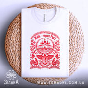 Футболка Київ серце Русі, біла, на плоскій поверхні, з червоним принтом, фон світло-сірий, принт з зображенням церкви та елементами українського орнаменту, розташована на плетеній основі.