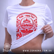 Футболка Київ серце Русі, біла, на плоскій поверхні, з червоним принтом, фон світло-сірий, принт з зображенням церкви та елементами українського орнаменту, з етикеткою.