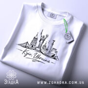 Футболка Київ Україна, біла, на плоскій поверхні, фон світло-сірий, принт з архітектурними елементами Києва, напис 'Kyiv, Ukraine' під малюнком, етикетка з зеленим шнурком.
