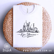 Футболка Київ Україна, біла, складена на плоскій поверхні, фон світло-сірий, принт з архітектурними елементами Києва, напис 'Kyiv, Ukraine' під малюнком, розташована на плетеній основі.