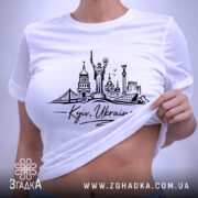 Футболка Київ Україна, біла, на плоскій поверхні, фон світло-сірий, принт з архітектурними елементами Києва, напис 'Kyiv, Ukraine' під малюнком, етикетка з зеленим шнурком.
