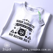 Футболка киця зайнята, біла, на столі, з верхнього ракурсу, фон світло-сірий, принт чорного кольору з зображенням кота та текстом, з біркою на зеленій стрічці.