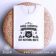 Футболка киця зайнята, біла, на столі, з верхнього ракурсу, фон натурального кольору, принт чорного кольору з зображенням кота та текстом.