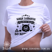 Футболка киця зайнята, біла, на столі, з верхнього ракурсу, фон світло-сірий, принт чорного кольору з зображенням кота та текстом, з біркою на зеленій стрічці.