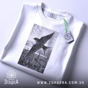Футболка ластівка над Києвом арт принт, біла футболка складена на поверхні, ракурс зверху, фон світло-сірий, принт зображує ластівку на фоні Київських будівель у чорно-білому стилі, з етикеткою зеленого кольору.