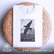 Футболка ластівка над Києвом арт принт, біла футболка складена на кошику, ракурс зверху, фон світло-сірий, принт зображує ластівку на фоні Київських будівель у чорно-білому стилі.