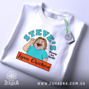 Футболка Лава Чікен, біла, на плоскій поверхні, з близьким ракурсом, фон світло-сірий, принт з персонажем, що махає рукою, текстом 'STEVE'S Tasty as hell!' та логотипом 'Lava Chicken', з етикеткою на комірці.