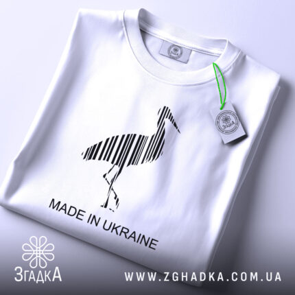Футболка Лелека, біла, знята зверху, на ній чорний принт лелеки з вертикальними смугами та написом 'MADE IN UKRAINE', етикетка з зеленим шнурком.