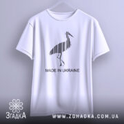 Футболка Лелека, біла, підвішена на вішалці, фон світло-фіолетовий, принт чорного лелеки з вертикальними смугами та написом 'MADE IN UKRAINE'.