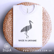 Футболка Лелека, біла, складена на плетеній основі, фон світлий, принт чорного лелеки з вертикальними смугами та написом 'MADE IN UKRAINE'.