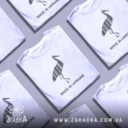 Футболка Лелека, біла, на моделі з коротким рукавом, фон світло-фіолетовий, принт у вигляді чорного лелеки з вертикальними смугами та написом 'MADE IN UKRAINE'.