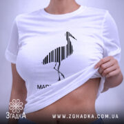 Футболка Лелека, біла, знята зверху, на ній чорний принт лелеки з вертикальними смугами та написом 'MADE IN UKRAINE', етикетка з зеленим шнурком.
