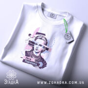 Футболка Леся Українка білого кольору, складена на поверхні, фон світло-сірий, принт з портретом Лесі Українки, прикрашений кольоровими елементами, з етикеткою на зеленій стрічці.