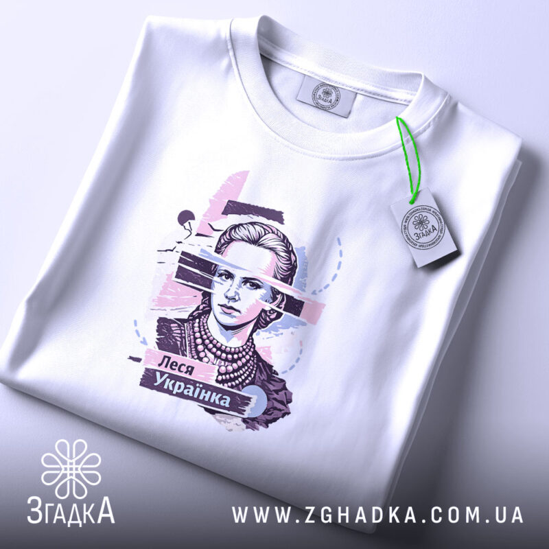 Футболка Леся Українка білого кольору, складена на поверхні, фон світло-сірий, принт з портретом Лесі Українки, прикрашений кольоровими елементами, з етикеткою на зеленій стрічці.