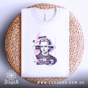 Футболка Леся Українка білого кольору, складена на плетеній кошику, фон світло-блакитний, принт з портретом Лесі Українки, оточений кольоровими абстрактними елементами.