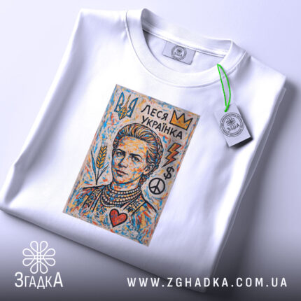 Футболка Леся Українка білого кольору, складена на столі. На фоні світло-сірий колір. Принт з портретом Лесі Українки, оточеним кольоровими елементами, такими як корона, серце, символи миру та грошей. На футболці підвішена етикетка з зеленим шнурком.