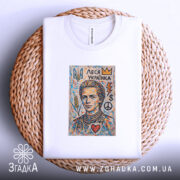 Футболка Леся Українка білого кольору, складена на плетеній кошику. На фоні світло-білий колір. Принт з портретом Лесі Українки, оточеним кольоровими елементами, такими як корона, серце, символи миру та грошей.