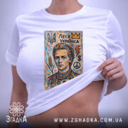 Футболка Леся Українка білого кольору, складена на столі. На фоні світло-сірий колір. Принт з портретом Лесі Українки, оточеним кольоровими елементами, такими як корона, серце, символи миру та грошей. На футболці підвішена етикетка з зеленим шнурком.