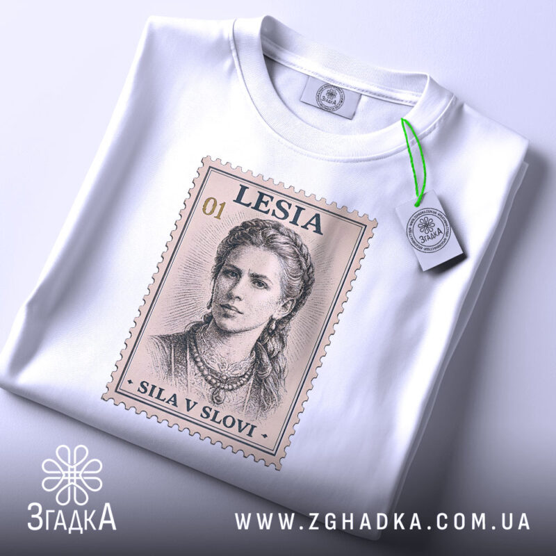 Футболка Леся Українка, біла, зображена зверху на плоскій поверхні, фон світло-сірий, принт у вигляді поштової марки з портретом Лесі Українки, текст 'SILA V SLOVI' під зображенням, на футболці прикріплена етикетка з зеленим шнурком.