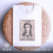 Футболка Леся Українка, біла, зображена на плоскій поверхні, фон світло-сірий, принт у вигляді поштової марки з портретом Лесі Українки, текст 'SILA V SLOVI' під зображенням, футболка лежить на плетеній основі.
