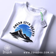 Футболка люблю бути зверху, біла, на столі, фон світло-фіолетовий, принт з горами та небом, текст чорного кольору, етикетка з зеленим шнурком.