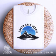 Футболка люблю бути зверху, біла, на кошику, фон світло-фіолетовий, принт з горами та небом, текст чорного кольору.