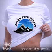 Футболка люблю бути зверху, біла, на столі, фон світло-фіолетовий, принт з горами та небом, текст чорного кольору, етикетка з зеленим шнурком.