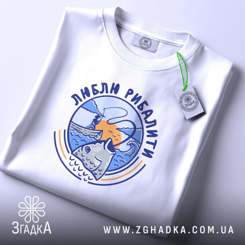 Футболка люблю рибалити, біла, складена на поверхні, фон світло-сірий, принт з рибою та рибалкою на фоні заходу сонця, з етикеткою.
