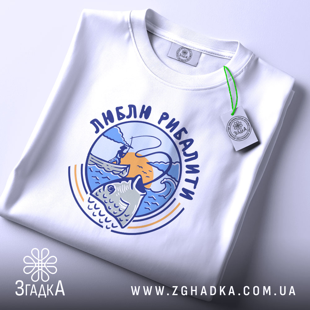 Футболка люблю рибалити, біла, складена на поверхні, фон світло-сірий, принт з рибою та рибалкою на фоні заходу сонця, з етикеткою.