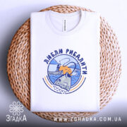 Футболка люблю рибалити, біла, складена на плетеній кошику, фон світло-сірий, принт з рибою та рибалкою на фоні заходу сонця.