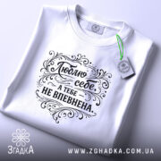 Футболка люблю себе, біла, складена на поверхні, фон світло-сірий, принт чорного кольору з написом 'Люблю себе, а тебе не впевнена' в декоративному стилі, з етикеткою зеленого кольору.