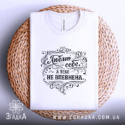 Футболка люблю себе, біла, складена на плетеній кошику, фон світло-сірий, принт чорного кольору з написом 'Люблю себе, а тебе не впевнена' в декоративному стилі.