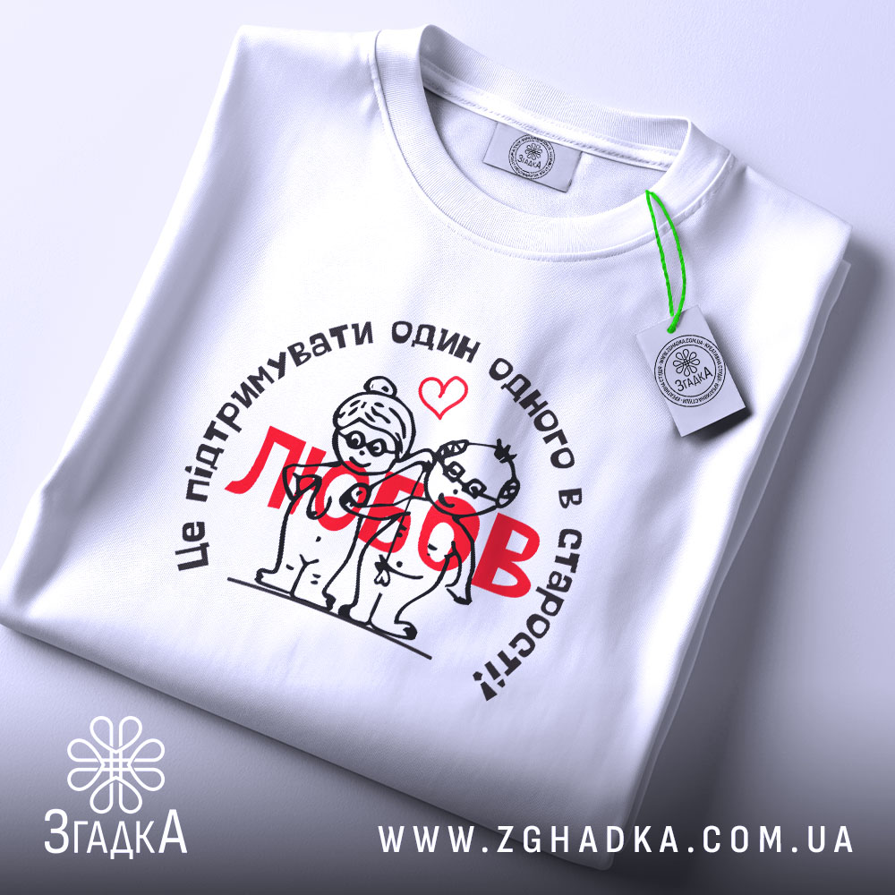 Футболка любов підтримка, біла, лежить на плоскій поверхні, фон світлий, принт з малюнком двох персонажів у старості, текст чорного кольору з червоним словом 'любов', на футболці присутня етикетка.