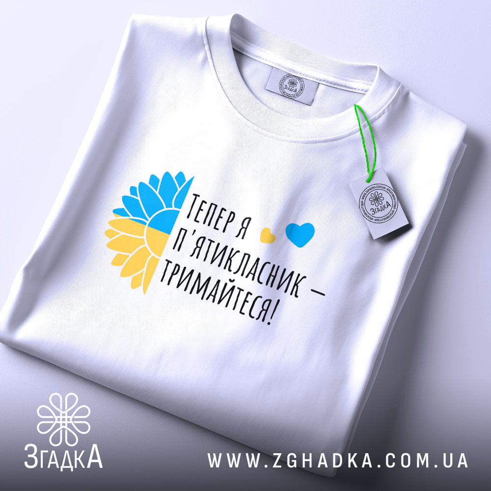 Футболка майбутнього пʼятикласника, білого кольору, зображена на плоскій поверхні, фон світло-сірий, принт містить текст "Тепер я п'ятикласник – тримайтеся!" з жовто-блакитними елементами, футболка має етикетку з зеленим шнурком.
