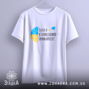 Футболка майбутнього пʼятикласника, білого кольору, підвішена на вішалці, фон світло-сірий, принт містить текст "Тепер я п'ятикласник – тримайтеся!" з кольоровими елементами у вигляді жовто-блакитного квіткового мотиву.