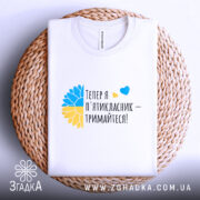 Футболка майбутнього пʼятикласника, білого кольору, розкладена на плоскій поверхні з плетеним елементом, фон світло-сірий, принт містить текст "Тепер я п'ятикласник – тримайтеся!" з жовто-блакитними елементами.