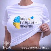 Футболка майбутнього пʼятикласника, білого кольору, зображена на плоскій поверхні, фон світло-сірий, принт містить текст "Тепер я п'ятикласник – тримайтеся!" з жовто-блакитними елементами, футболка має етикетку з зеленим шнурком.