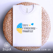 Футболка майбутньої пʼятикласниці білого кольору, складена на плетеній кошику. Принт з жовто-блакитним соняшником та текстом "Тепер я п'ятікласниця – тримайтеся!" добре видимий. Фон натуральний, що підкреслює текстуру кошика.