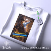 Футболка мама головнокомандувачка, білого кольору, зображена на плоскій поверхні, принт з жінкою в халаті на коні, що вказує вперед, з яскравими кольорами та текстом, на етикетці видно логотип бренду.