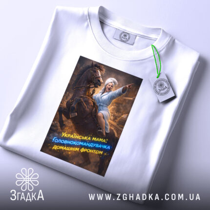 Футболка мама головнокомандувачка, білого кольору, зображена на плоскій поверхні, принт з жінкою в халаті на коні, що вказує вперед, з яскравими кольорами та текстом, на етикетці видно логотип бренду.